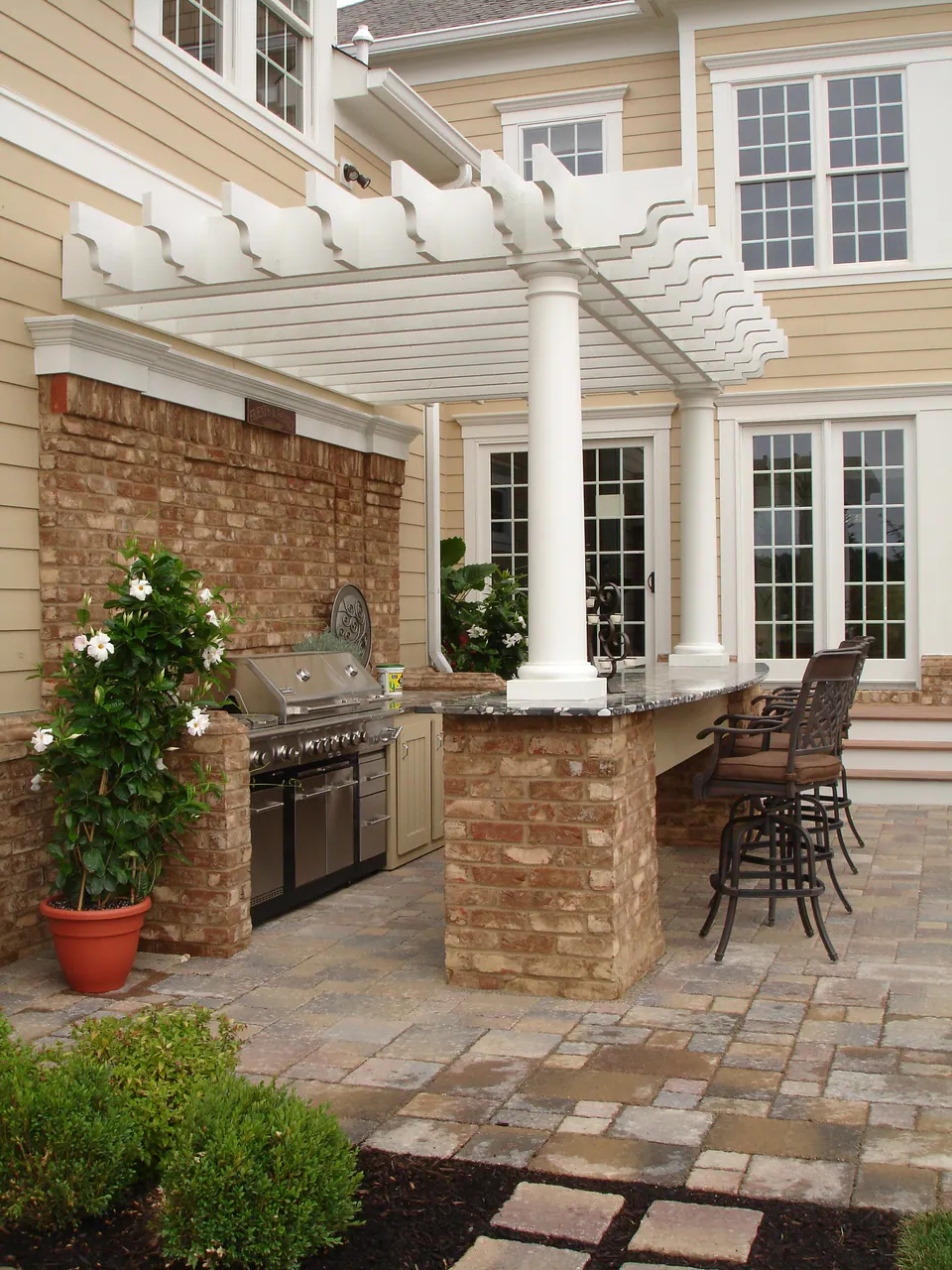 Brick and stone outdoor kitchen with grill and bar seating under a white pergola beside the house